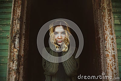 A beautiful blonde girl in a wool sweater stands alone in the dark doorway of an abandoned wooden house. Evil and mysticism Stock Photo
