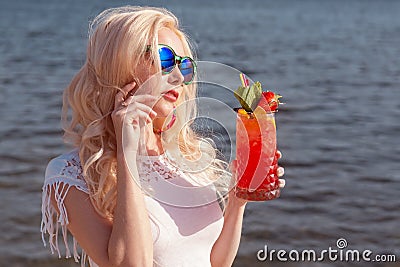 Beautiful blonde girl with a red beautiful cocktail in her hands by the sea / river Stock Photo