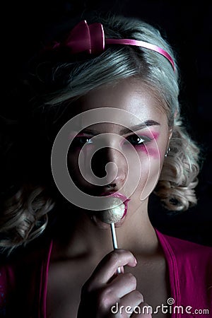 Beautiful blonde girl with two pigtails, with creative doll make-up: pink glossy lips, wearing pink skeleton dress licking lollipo Stock Photo