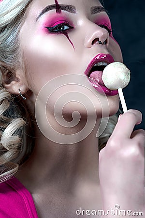 Beautiful blonde girl with two pigtails, with creative doll make-up: pink glossy lips, wearing pink skeleton dress licking lollipo Stock Photo