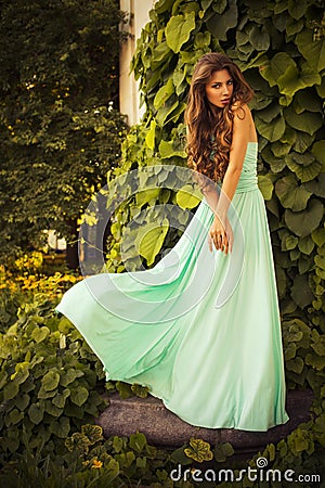 Beautiful blonde with a long curly hair in a long evening dress in static outdoors near retro vintage building in summer sunset Stock Photo