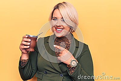 Beautiful blonde plus size woman drinking mate infusion beverage over yellow background smiling happy pointing with hand and Stock Photo