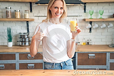 Beautiful blonde shows thumb up looking at camera while holding orange juice in her hand and smiling. Health Care And Beauty Stock Photo