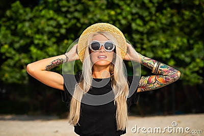 Beautiful blonde is smiling and looking at the morning sea. Holds a hat with his hands with tattoos. Travel to Thailand. Stock Photo