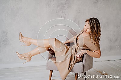 A blonde woman in a beige dress poses on a gray chair in a photo studio. Stock Photo