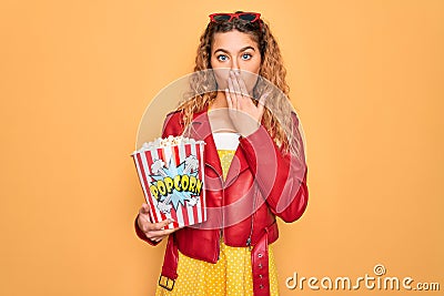 Beautiful blonde woman with blue eyes eating salty popcorn snack over yellow background cover mouth with hand shocked with shame Stock Photo