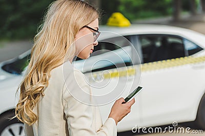 beautiful blonde woman in eyeglasses using smartphone while standing near taxi Stock Photo