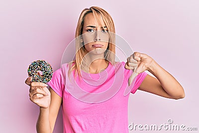 Beautiful blonde woman holding donut with angry face, negative sign showing dislike with thumbs down, rejection concept Stock Photo