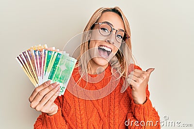 Beautiful blonde woman holding norwegian krone banknotes pointing thumb up to the side smiling happy with open mouth Stock Photo