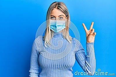 Beautiful blonde woman wearing covid-19 medical mask smiling with happy face winking at the camera doing victory sign Stock Photo