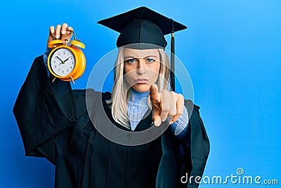 Beautiful blonde woman wearing graduation cap and ceremony robe holding alarm clock pointing with finger to the camera and to you, Stock Photo