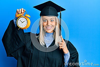 Beautiful blonde woman wearing graduation cap and ceremony robe holding alarm clock surprised with an idea or question pointing Stock Photo