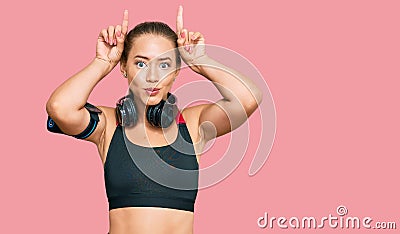 Beautiful blonde woman wearing gym clothes and using headphones doing funny gesture with finger over head as bull horns Stock Photo