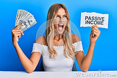 Beautiful blonde young woman holding dollars and passive income text angry and mad screaming frustrated and furious, shouting with Stock Photo