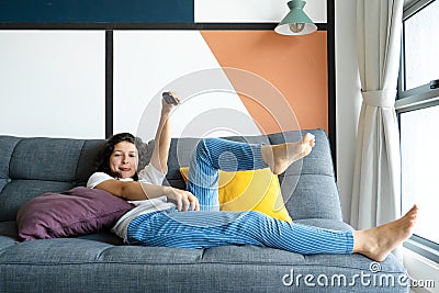 Beautiful bored brunette girl on the couch in a stylish interior is watching TV. Switches the TV channel with a remote control in Stock Photo