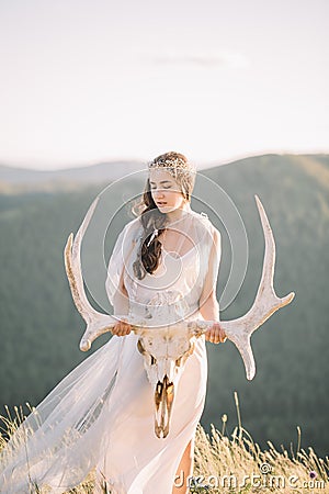Beautiful bride brunette girl in white long dress is made of tulle looks down, keeps on hand a deer skull with horns decoration on Stock Photo