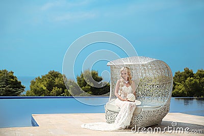 Beautiful Bride Portrait holding wedding bouquet posing in formal prom lace mermaid dress. Beautiful blond sitting on beach sofa Stock Photo