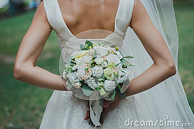 Beautiful bride in satin lace dress and veil is holding bouquet behind her back. Roses, eustoma and eucalyptus elegant flowers. Stock Photo