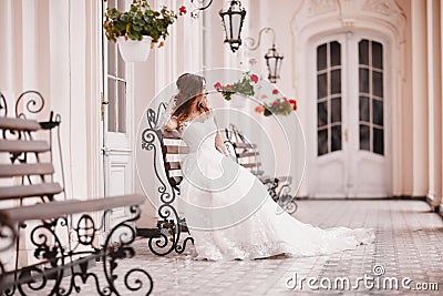Beautiful bride is sitting on wooden bench in old city. young woman with long hair in lace satin wedding dress in Stock Photo
