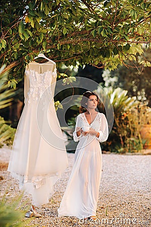 A beautiful bride stands next to a wedding dress with a Cup of tea in a boudoir outfit next to a Villa in Italy.morning of the Stock Photo