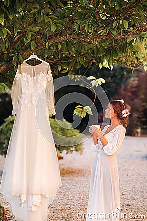 A beautiful bride stands next to a wedding dress with a Cup of tea in a boudoir outfit next to a Villa in Italy.morning of the Stock Photo