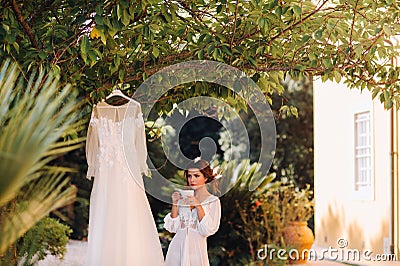 A beautiful bride stands next to a wedding dress with a Cup of tea in a boudoir outfit next to a Villa in Italy.morning of the Stock Photo