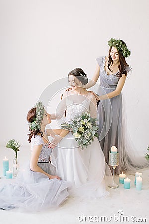 Beautiful bride in a white wedding dress holding bouquet and sitting on the vintage chair with bridesmaids in blue grey Stock Photo