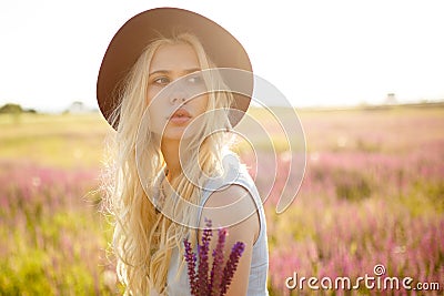 Beuty portrait of gorgeous blonde girl wearing in hat posing outside, isolated on a floral field, on sunset background. Stock Photo