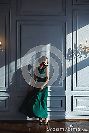 A beautiful brunette girl with modish hairstyle, in green evening dress, dancing near the blue wall with sunlight and Stock Photo