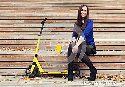 Beautiful brunette girl sitting on wooden steps with a glass and donuts. Yellow scooter stands next Stock Photo