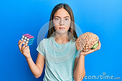 Beautiful brunette little girl playing colorful puzzle cube intelligence game and holding brain relaxed with serious expression on Stock Photo