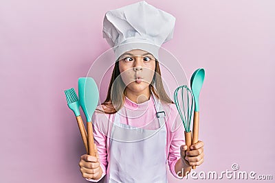 Beautiful brunette little girl wearing professional cook apron holding cooking tools making fish face with mouth and squinting Stock Photo