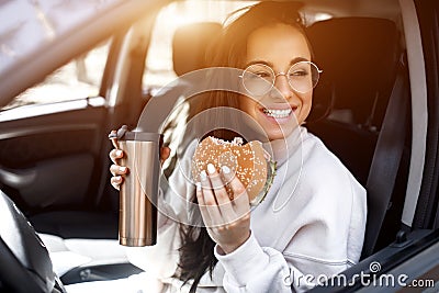 Beautiful brunette model has lunch in a car. Woman eats a hamburger and drinks coffee or tea from a thermos. Food on the Stock Photo
