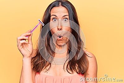 Beautiful brunette woman asking for depilation holding shaver razor over yellow background scared and amazed with open mouth for Stock Photo