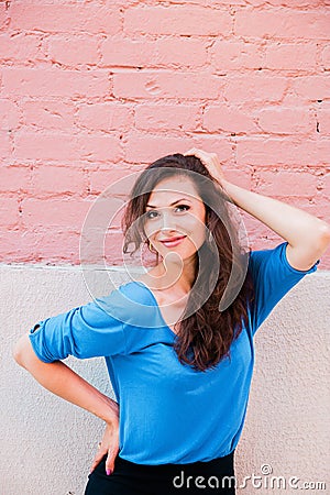 A beautiful brunette woman in a blue blouse posing against a textured pink brick wall. Portrait of a young woman Stock Photo