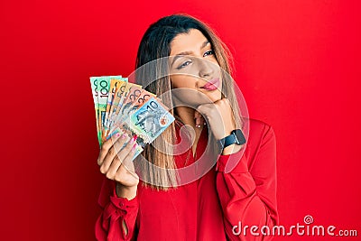 Beautiful brunette woman holding australian dollars with hand on chin thinking about question, pensive expression Stock Photo