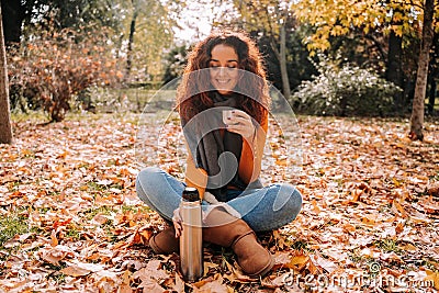 A beautiful brunette woman sitting on the ground surrounded by leaves while enjoying a coffee in the park. She is putting the Stock Photo