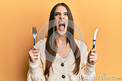 Beautiful brunette young woman holding fork and knife ready to eat angry and mad screaming frustrated and furious, shouting with Stock Photo