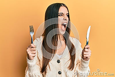 Beautiful brunette young woman holding fork and knife ready to eat angry and mad screaming frustrated and furious, shouting with Stock Photo