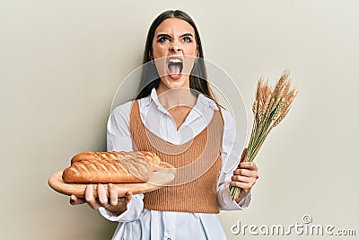Beautiful brunette young woman holding homemade bread and spike wheat angry and mad screaming frustrated and furious, shouting Stock Photo