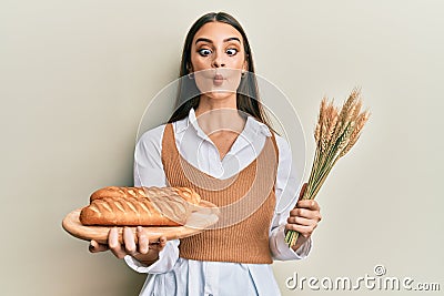 Beautiful brunette young woman holding homemade bread and spike wheat making fish face with mouth and squinting eyes, crazy and Stock Photo