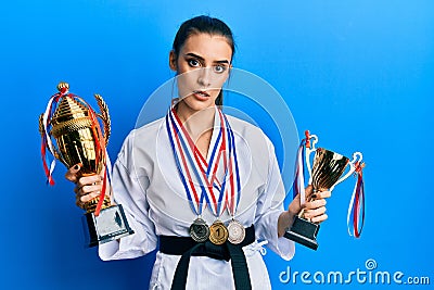 Beautiful brunette young woman wearing karate fighter uniform and medals holding trophy clueless and confused expression Stock Photo