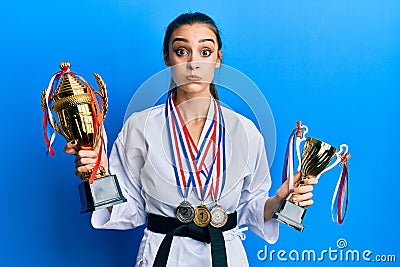 Beautiful brunette young woman wearing karate fighter uniform and medals holding trophy puffing cheeks with funny face Stock Photo