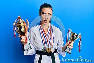 Beautiful brunette young woman wearing karate fighter uniform and medals holding trophy in shock face, looking skeptical and Stock Photo