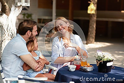 Beautiful blonde laughing and looking at young beardy dad with his daughter in arms, sitting in his lap Stock Photo