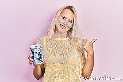 Beautiful caucasian blonde woman holding piggy bank dollars moneybox pointing thumb up to the side smiling happy with open mouth Stock Photo