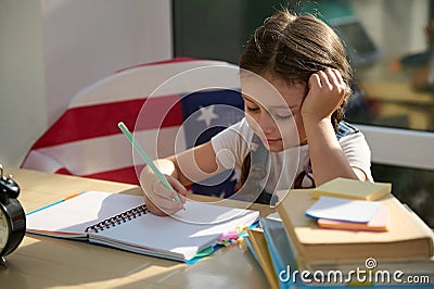 Beautiful Caucasian kid, schoolgirl, first grader sitting on a chair with American flag, using pencil learns writing Stock Photo