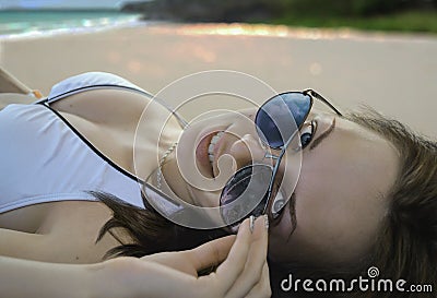 A beautiful Caucasian female in white bikini lying on a sandy beach by the sea, smiling and holding her sunglasses. A Stock Photo