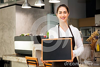 Beautiful Caucasian woman in barista apron holding empty blackboard sign inside coffee shop - ready to insert text Stock Photo
