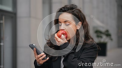 Beautiful Caucasian woman walking outdoors, wearing black jacket and white teeshirt, scrolling her smartphone and eating Stock Photo